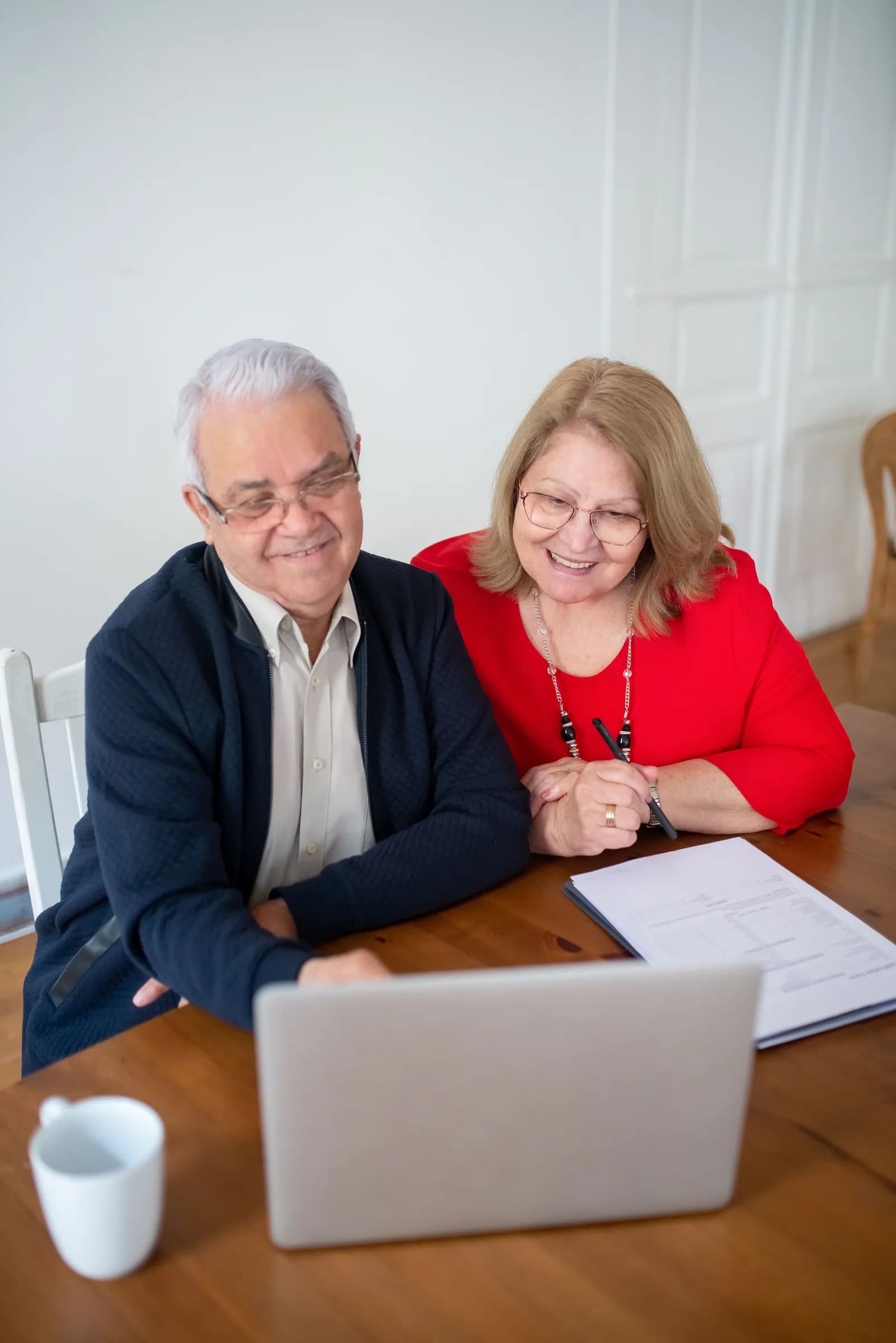 A retired couple sitting side by side at a dining table, smiling together at a laptop with a printed form beside it.