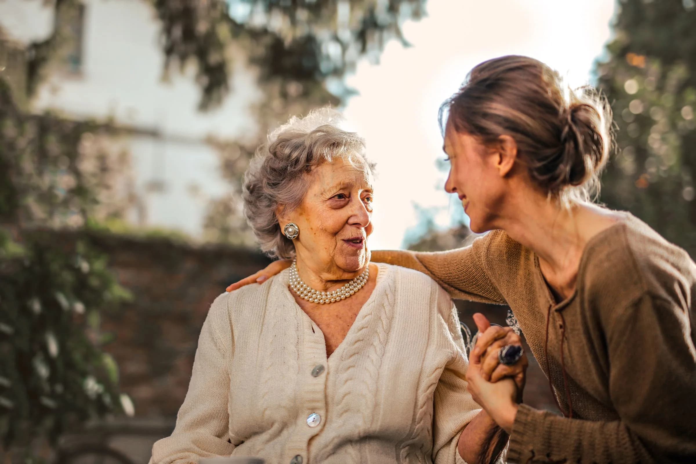 An older woman and a younger woman standing close together in a garden, talking and smiling.