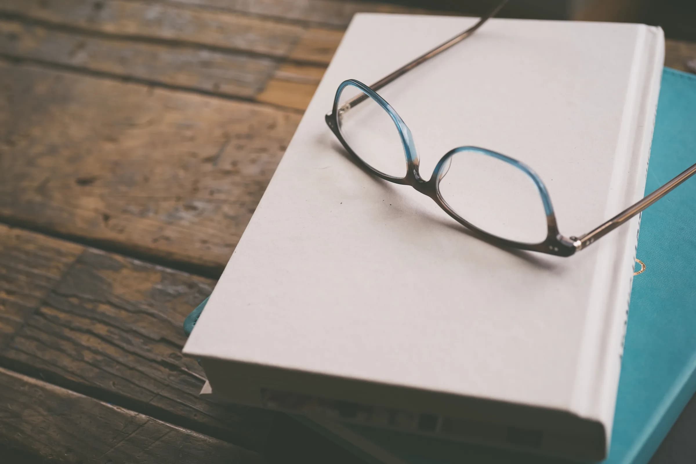 A pair of reading glasses resting on a stack of closed hardcover books on a rustic wooden table.