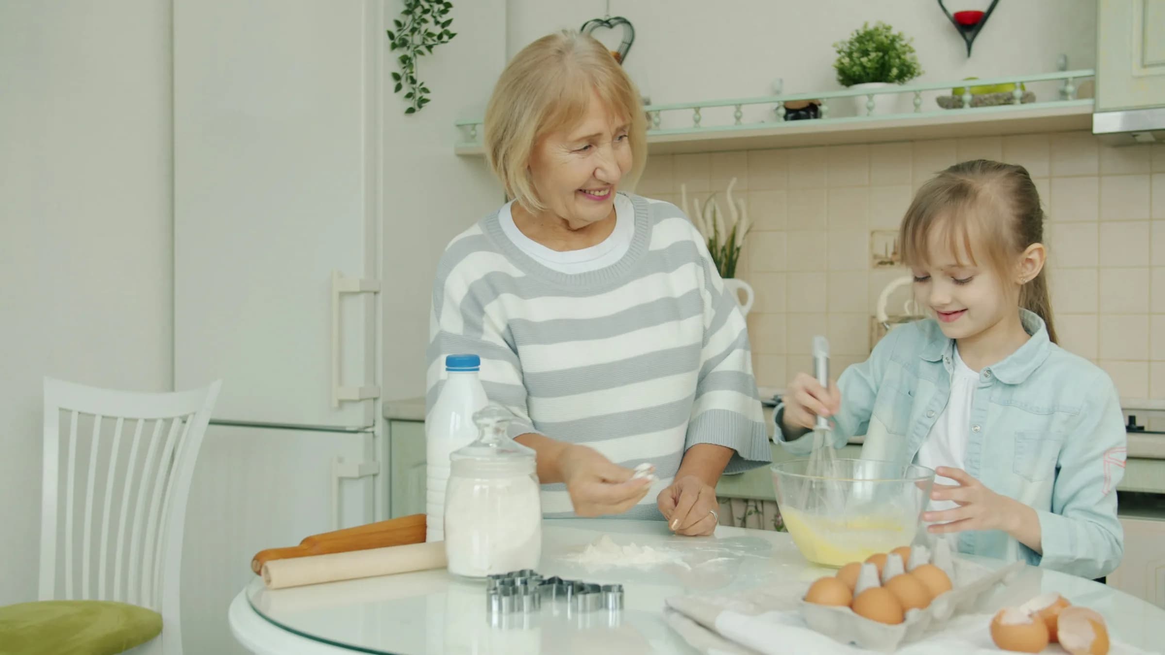A grandmother and granddaughter baking together at a kitchen table, smiling as the girl whisks a bowl of batter.