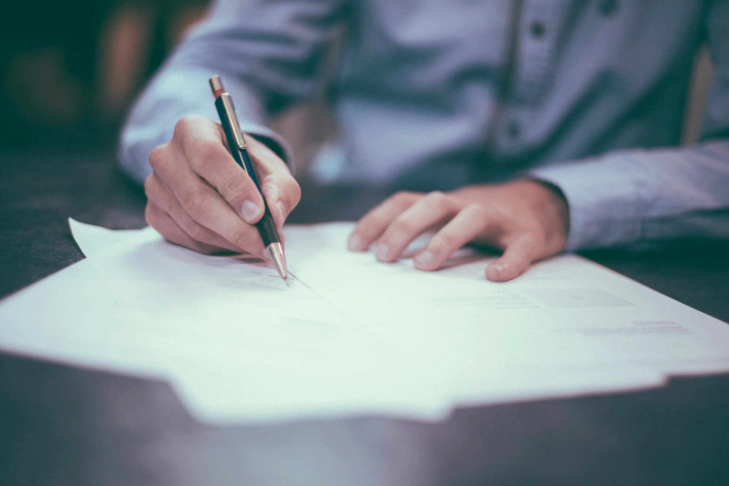 A close view of two hands at a desk, one steadying a printed page while the other signs it with a pen.